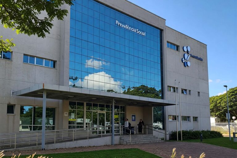 Meu INSS: Prédio moderno da Previdência Social com fachada espelhada, passarela de acesso coberta, área externa com gramado e caminho asfaltado, em um dia ensolarado. A fachada exibe a inscrição "Previdência Social". Gerada por AI.