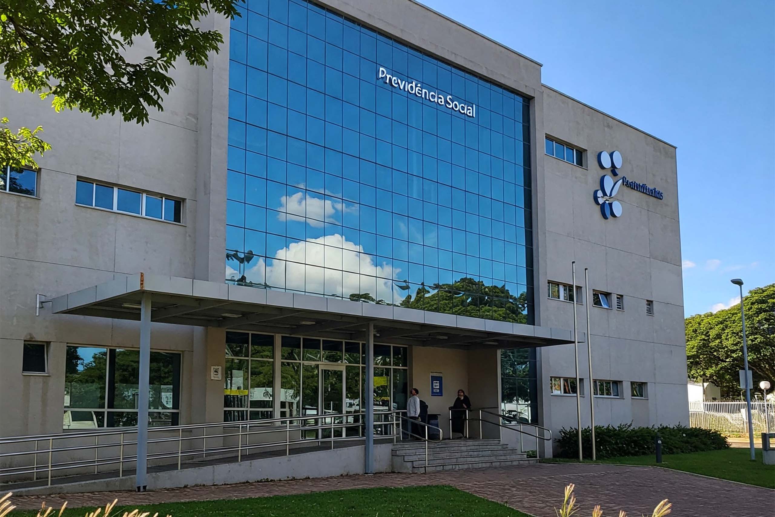 Meu INSS: Prédio moderno da Previdência Social com fachada espelhada, passarela de acesso coberta, área externa com gramado e caminho asfaltado, em um dia ensolarado. A fachada exibe a inscrição "Previdência Social". Gerada por AI.