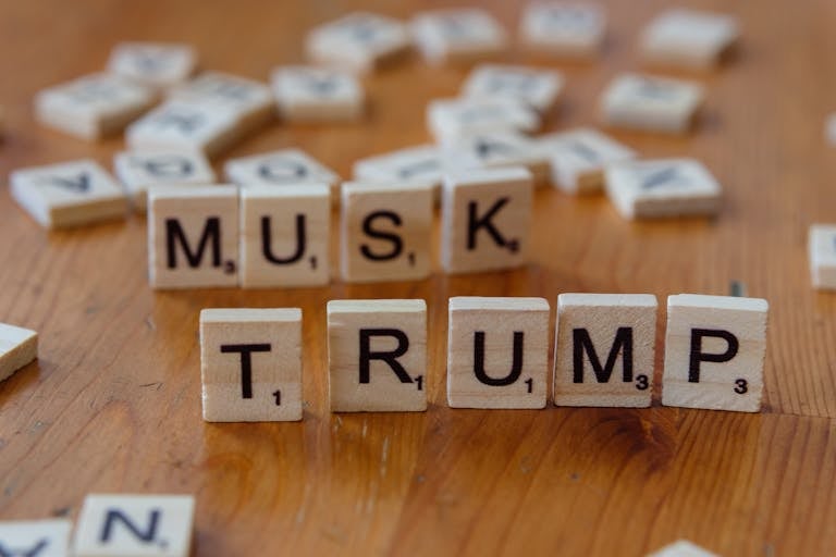 Peças de Scrabble formando os nomes de Trump e Elon Musk sobre uma mesa de madeira.